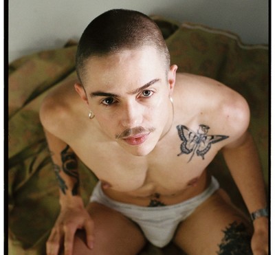 Oscar, a trans man, looks up at the camera. He's shirtless, has tattoos, and a moustache.
